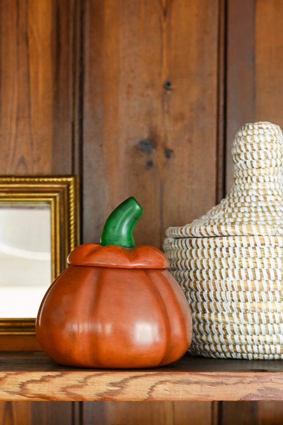 An orange pumpkin-shaped ceramic container with a green stem sits on a wooden shelf next to a woven basket and a gold picture frame against a dark wood paneled wall.