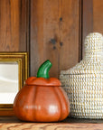 An orange pumpkin-shaped ceramic container with a green stem sits on a wooden shelf next to a woven basket and a gold picture frame against a dark wood paneled wall.