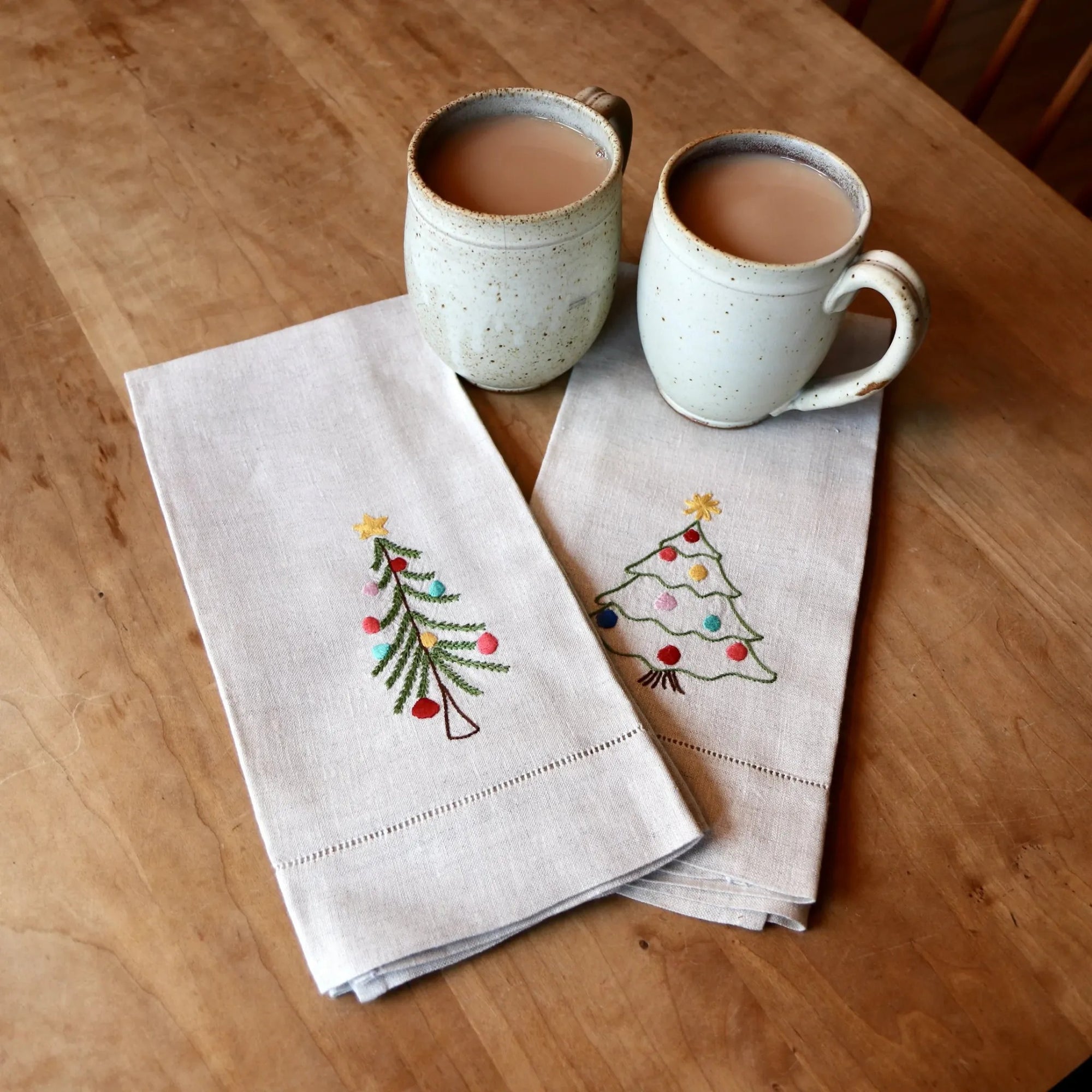 Two ceramic mugs with a beverage on a wooden table, accompanied by two embroidered napkins with Christmas tree designs.
