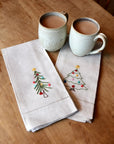 Two ceramic mugs with a beverage on a wooden table, accompanied by two embroidered napkins with Christmas tree designs.