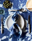 Table setting with a blue floral tablecloth, white plates, silverware, and a blue napkin.