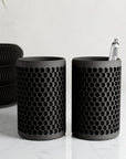 Two black honeycomb-patterned cylindrical containers on a light surface.
