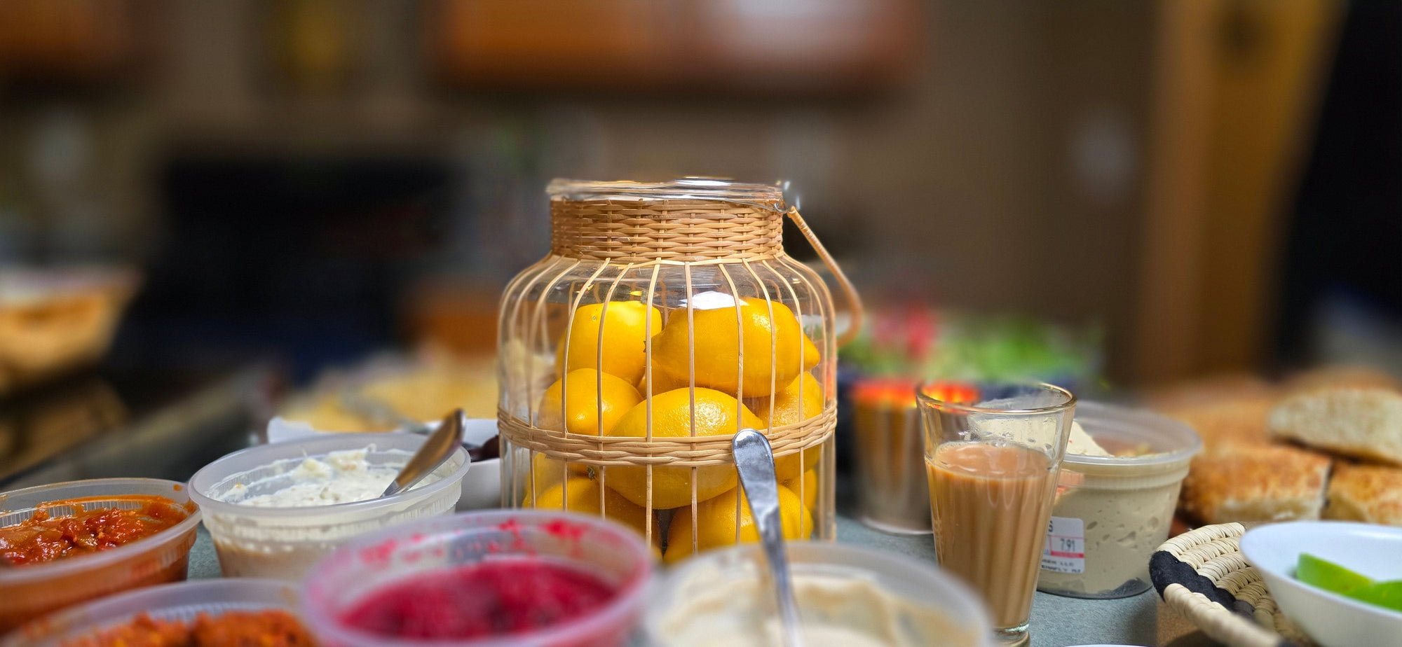 Basket of lemons on a table inside a wicker and glass jar with various food items in the background
