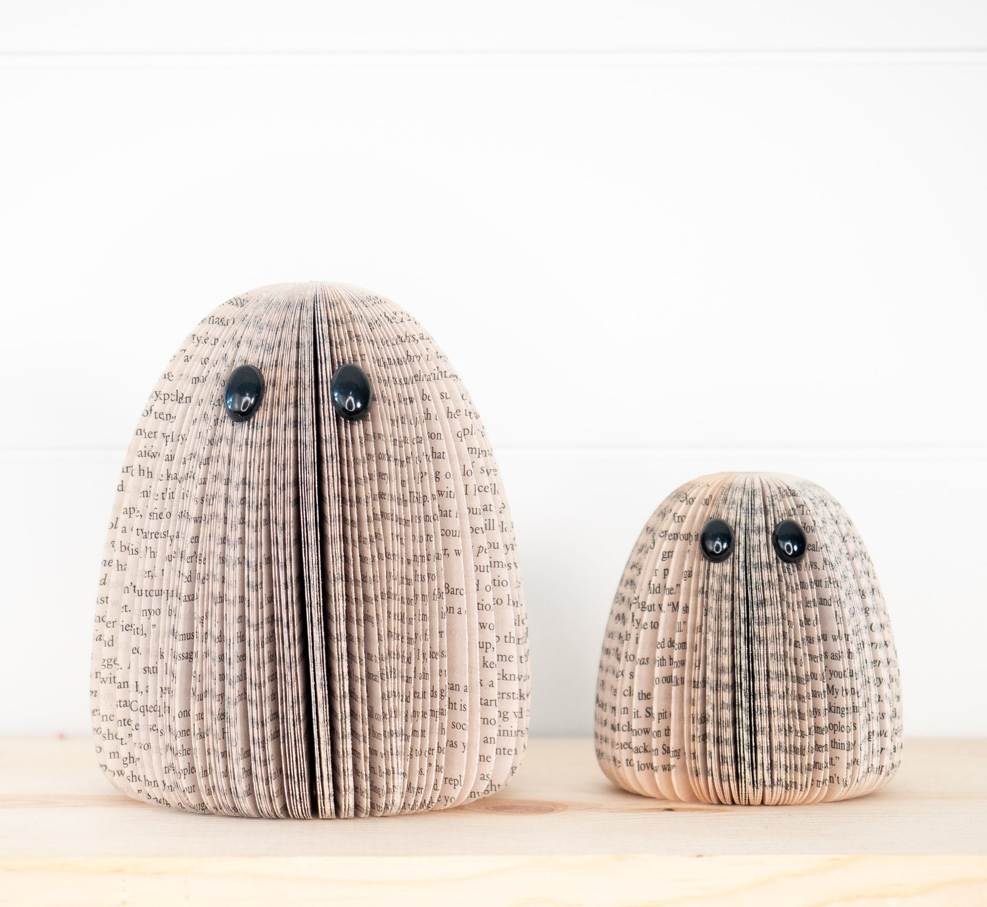 Two decorative book-folded ghost sculptures in different sizes, made from beige and brown book pages with black text, featuring black button eyes, displayed on a light wooden surface against a white paneled wall.