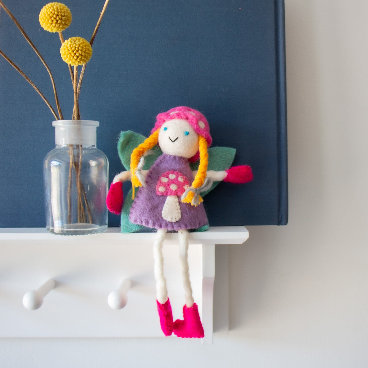 Blonde felt tooth fairy in a pink mushroom bonnet sitting on a white shelf beside a vase.