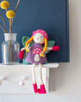 Blonde felt tooth fairy in a pink mushroom bonnet sitting on a white shelf beside a vase.