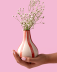 Hand holding a red striped ceramic vase with dried flowers against a pink background