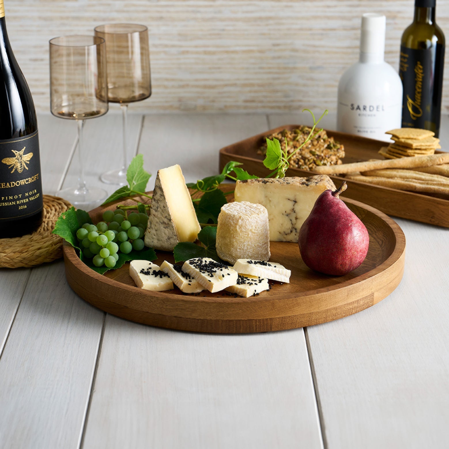 Wooden tray with assorted cheeses, fruits, and a bottle of wine on a light wooden surface.