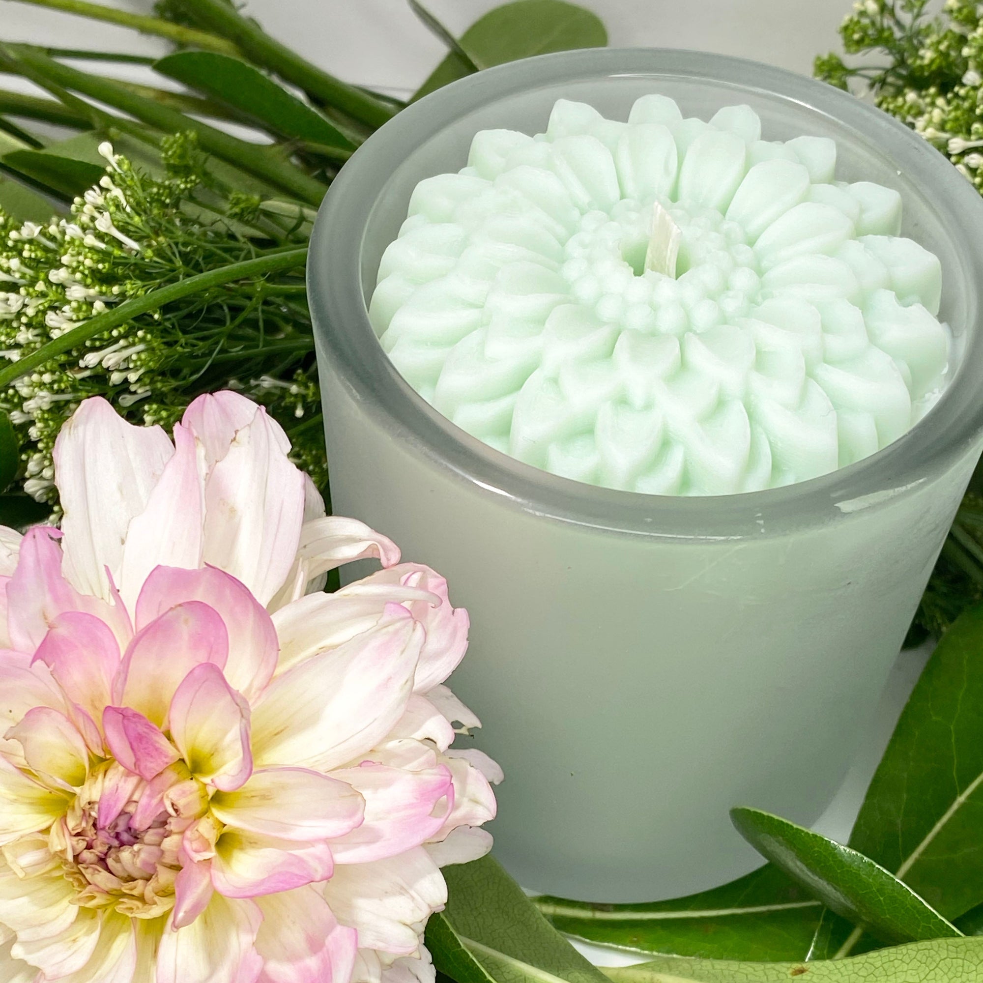 Blue flower candle in a glass container with a pink flower and green leaves in the background