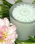 Blue flower candle in a glass container with a pink flower and green leaves in the background