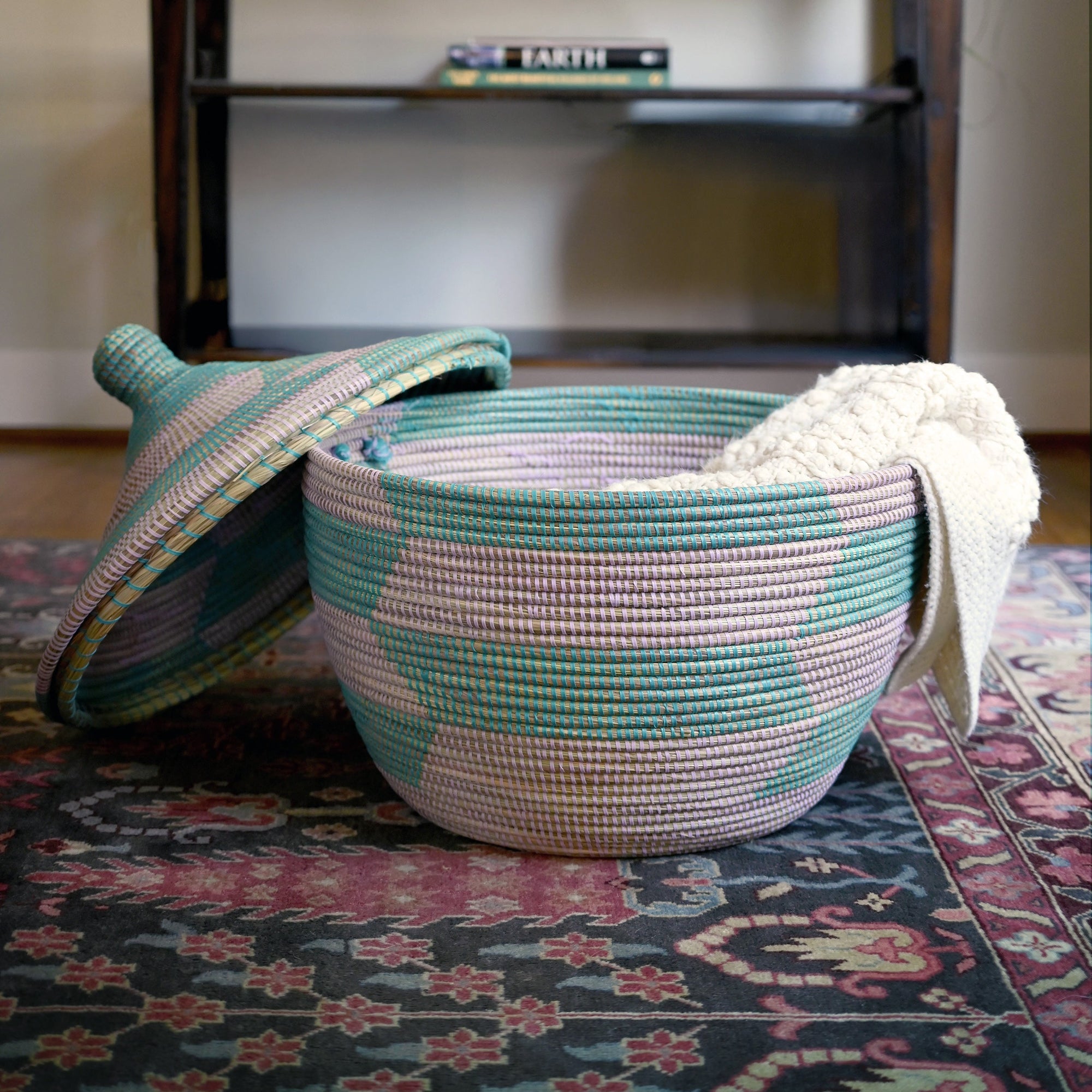 Woven basket with a patterned lid on a decorative rug, with a shelf in the background.