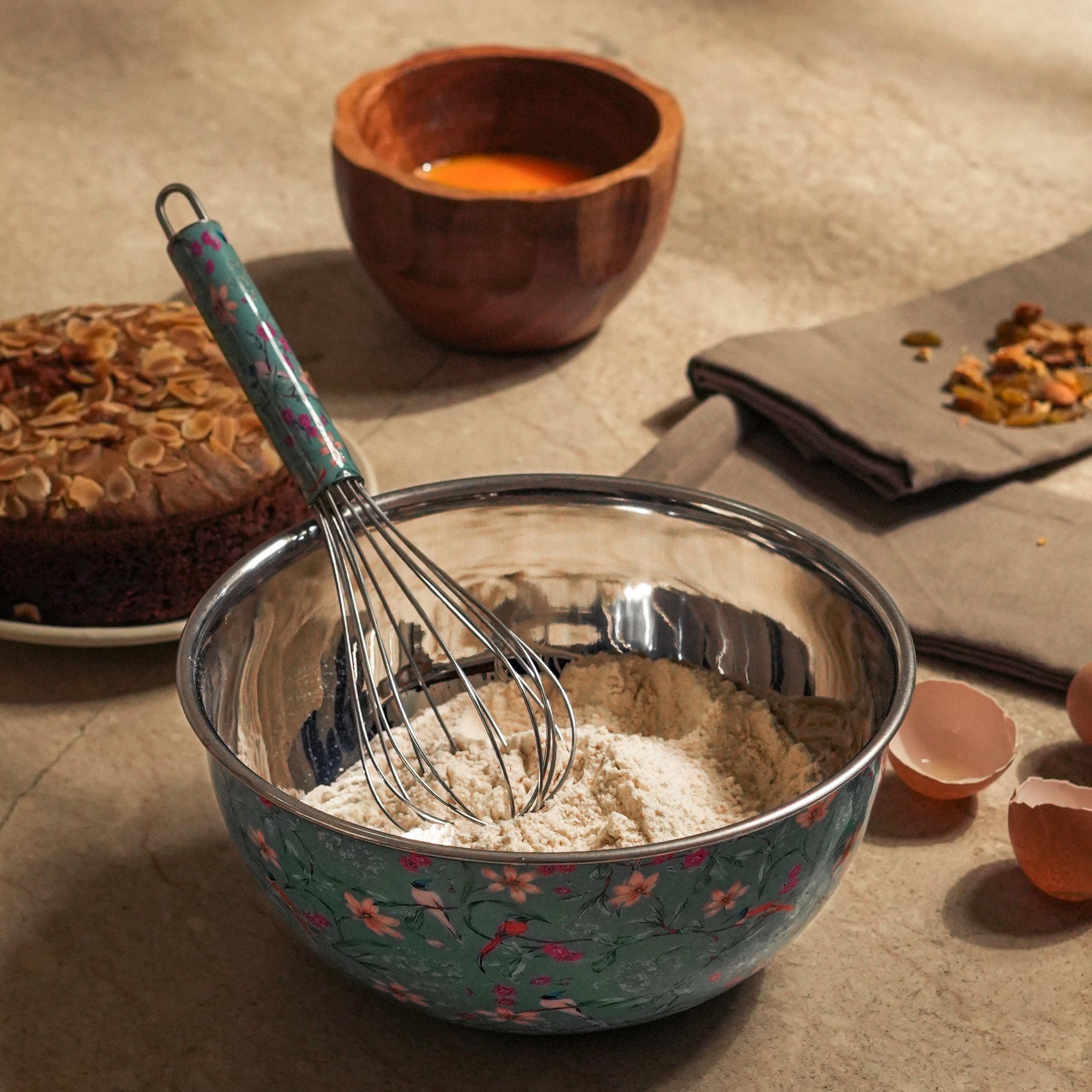 Metal mixing bowl with flour and a whisk on a rustic surface with a wooden bowl in the background.