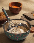 Metal mixing bowl with flour and a whisk on a rustic surface with a wooden bowl in the background.