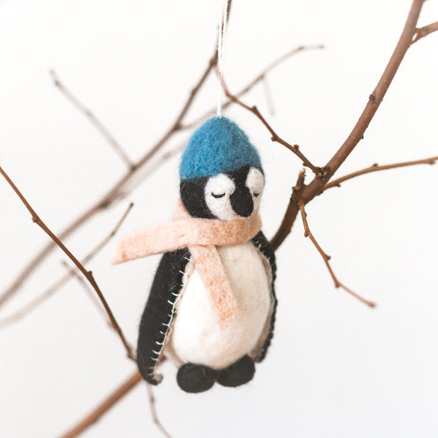 Felt penguin ornament with a blue hat and pink scarf hanging on bare branches.